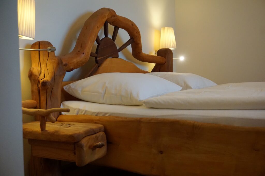 Ein gemütlich wirkendes Schlafzimmer mit einem einzigartigen, aus natürlichem Holz gefertigten Bettgestell und weißen Bettdecken. An jeder Seite des Bettes befindet sich eine kleine Nachtlampe.