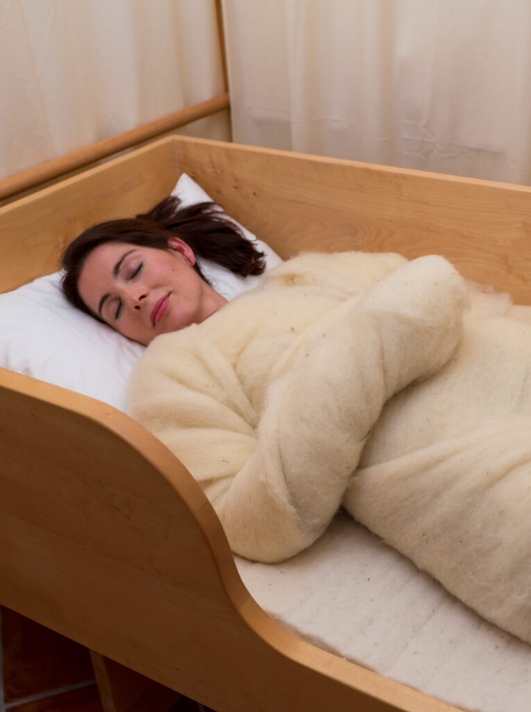 Frau schläft friedlich in einem Holzbett, zugedeckt mit einer dicken, hellen Decke.