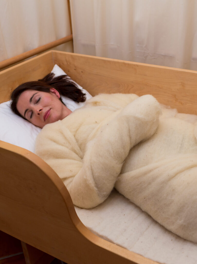 Eine Frau schläft friedlich in einem Holzbett, zugedeckt mit einer hellen Wolldecke.