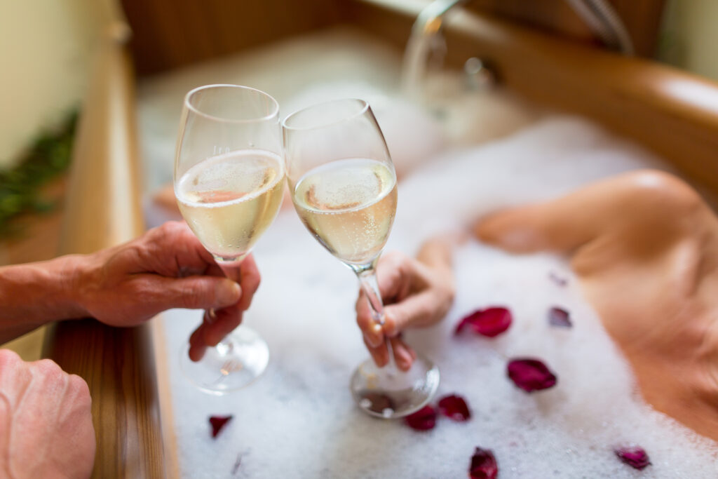 Zwei Personen stoßen in einer Badewanne mit Schaum und Rosenblättern mit Sektgläsern an.