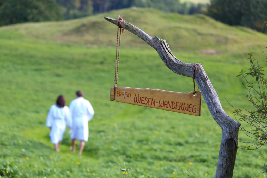 Ein Paar in weißen Bademänteln spaziert auf einer Wiese entlang, vorbei an einem Holzschild mit der Aufschrift "Barfuß-Wiesen-Wanderweg".