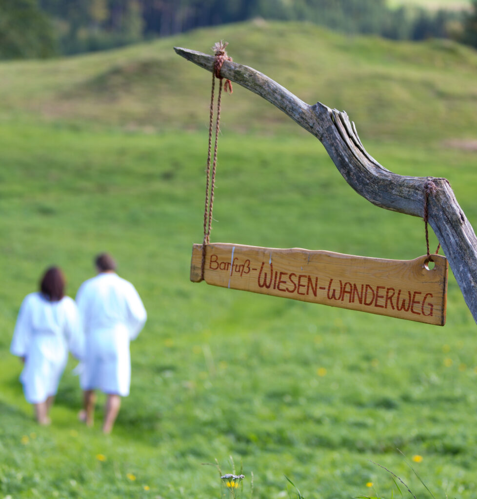 "Holzschild mit der Aufschrift 'Barfuß-WIESEN-WANDERWEG' hängt an einem Ast, im Hintergrund gehen zwei Personen in weißen Bademänteln auf einer Wiese."