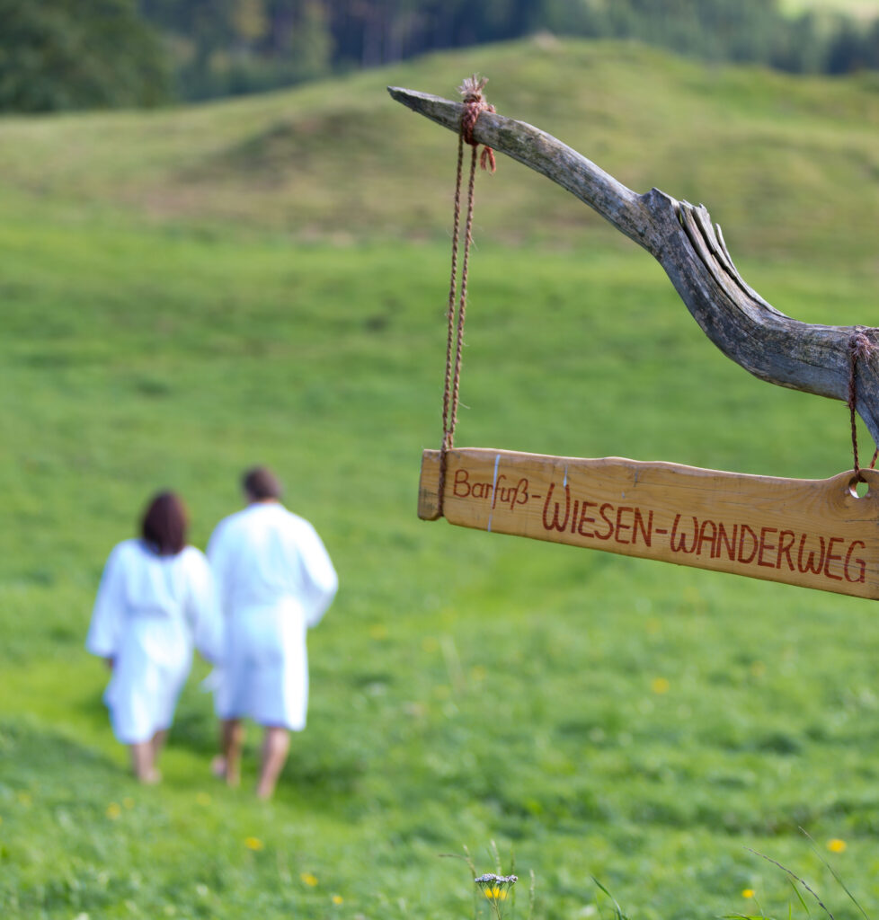 "Zwei Personen in weißen Mänteln gehen auf einer Wiese spazieren, vorne ein Schild mit der Aufschrift 'Barfuß-Wiesen-Wanderweg', aufgehängt an einem gebogenen Ast."
