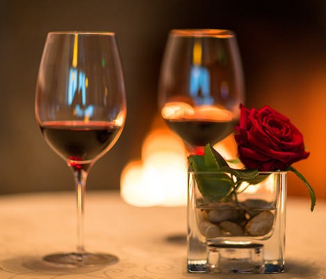 Zwei Weingläser mit Rotwein auf einem Tisch, neben ihnen steht ein kleines Glas mit einer roten Rose, unscharfer Hintergrund mit warmem Licht.