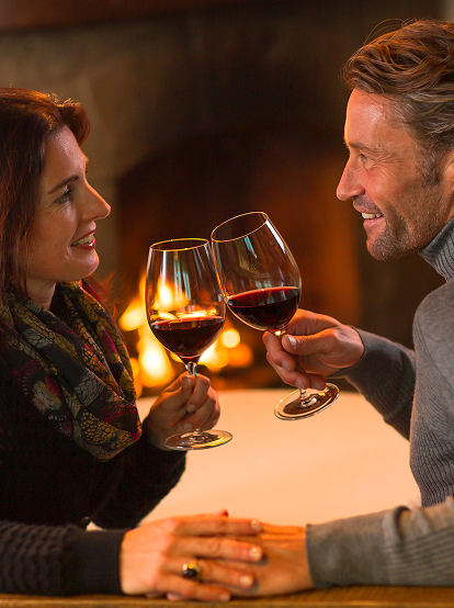 Ein Mann und eine Frau stoßen mit Rotweingläsern an einem gemütlichen Kaminfeuer an.