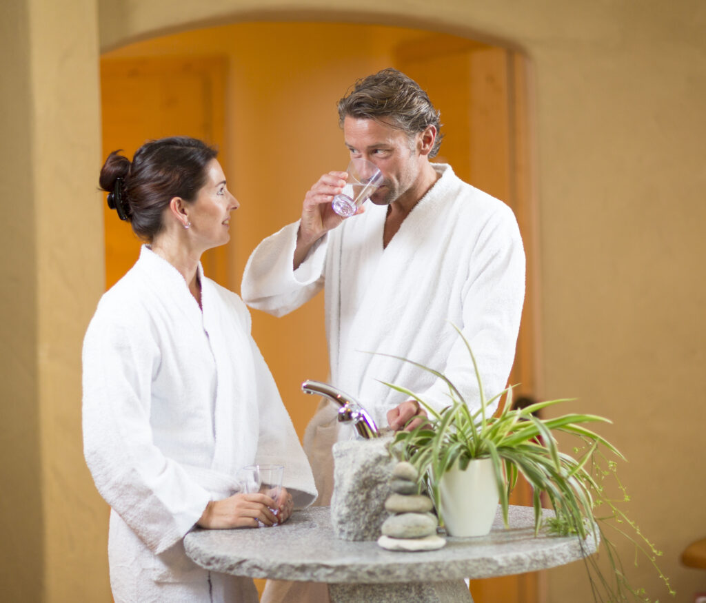 Ein Mann und eine Frau in weißen Bademänteln stehen in einem Spa und trinken Wasser.