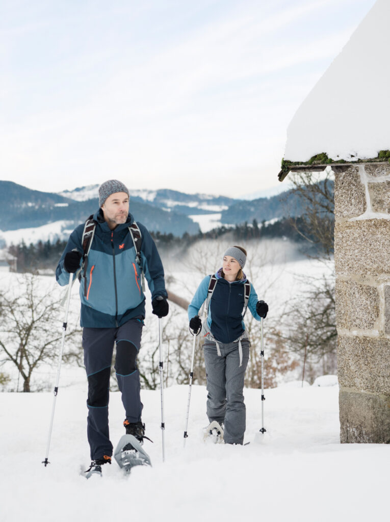 Ein Mann und eine Frau gehen mit Schneeschuhen in einer schneebedeckten Landschaft neben einem Haus spazieren.