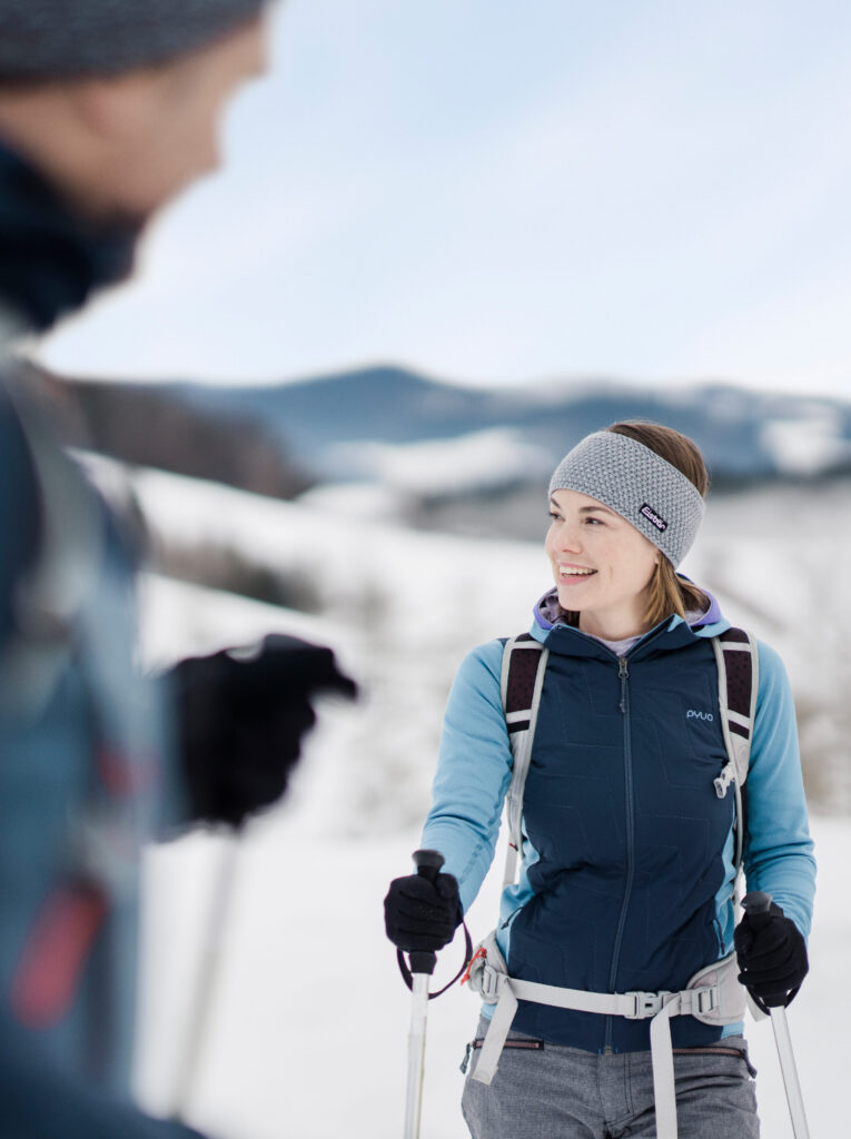 Eine junge Frau mit Rucksack und Winterkleidung steht im Schnee und unterhält sich mit einer außerhalb des Bildes befindlichen Person, während sie Langlaufstöcke hält. Im Hintergrund sind schneebedeckte Berge zu sehen.