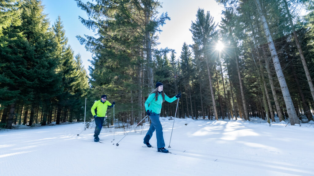 Zwei Personen beim Langlaufen in einem verschneiten Wald, Sonnenstrahlen durchbrechen die Baumwipfel.