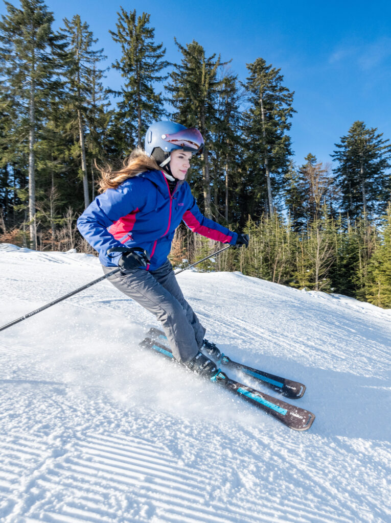 Junge Frau beim Skifahren auf einer sonnigen, verschneiten Piste mit Bäumen im Hintergrund.