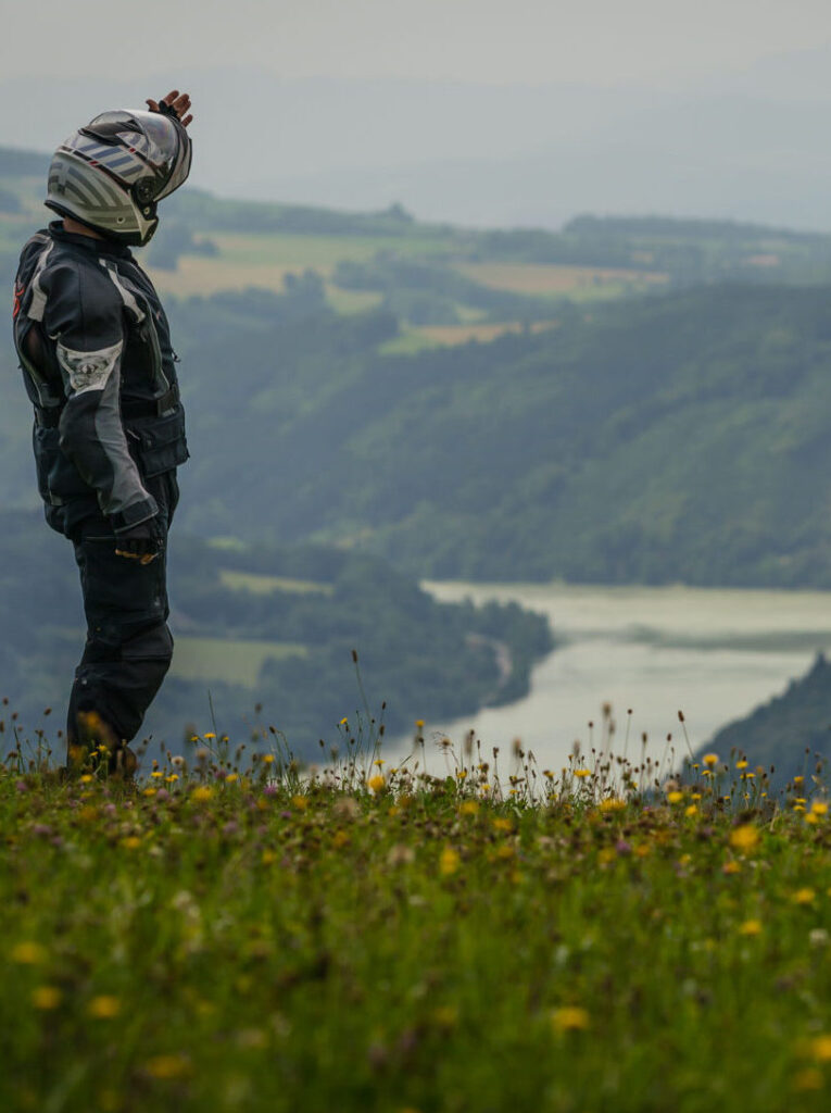 Motorradfahrer in Schutzkleidung steht auf einer Wiese und blickt auf einen Fluss im Tal.