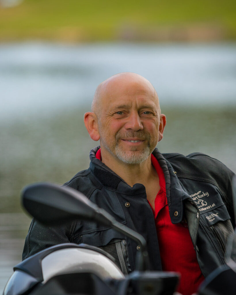 Ein lächelnder Mann mittleren Alters mit Glatze, bekleidet mit einer schwarzen Lederjacke und rotem Hemd, sitzt neben einem Motorrad vor einem unscharfen Hintergrund mit Wasser.