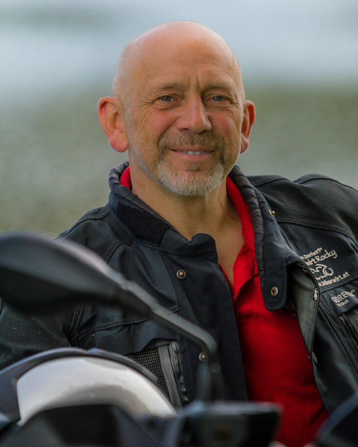 Ein lächelnder, kahlköpfiger Mann mittleren Alters in einer schwarzen Lederjacke und einem roten Shirt, der neben einem Motorrad sitzt.