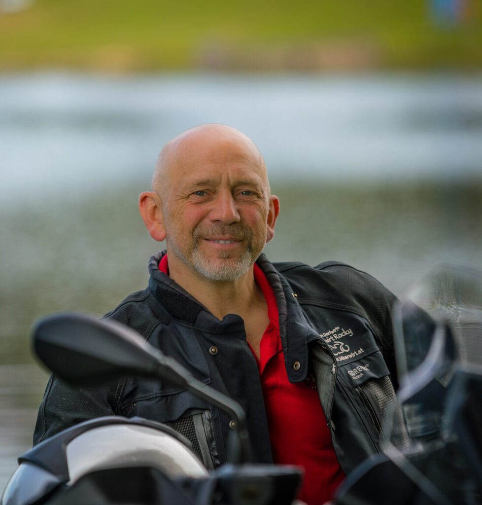 Ein lächelnder Mann mit Glatze trägt eine Lederjacke und sitzt neben einem Motorrad vor einem unscharfen natürlichen Hintergrund.