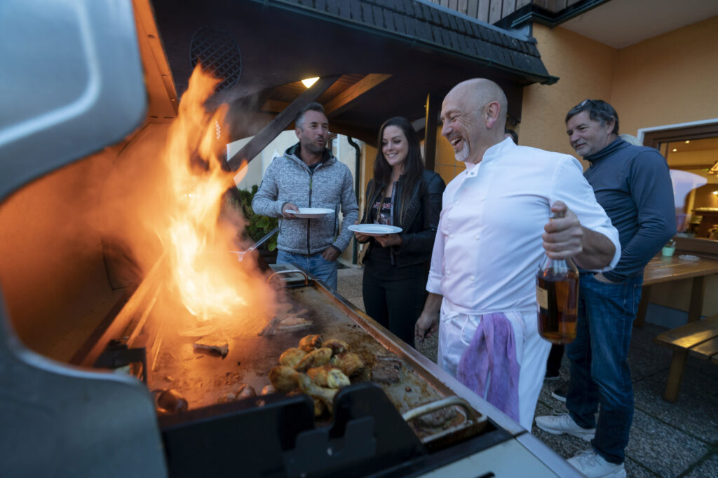 Eine Gruppe von Freunden genießt ein Grillfest im Freien; ein Mann bedient den Grill, auf dem Flammen hochschlagen, während andere mit Tellern und Getränken um ihn herumstehen.