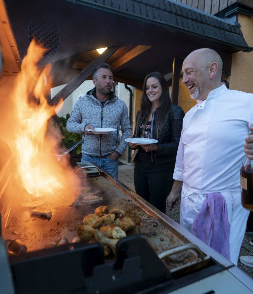 Drei Personen, darunter ein Koch in Kochkleidung, amüsieren sich an einem Grill mit hoher Flamme im Freien während der Abenddämmerung.