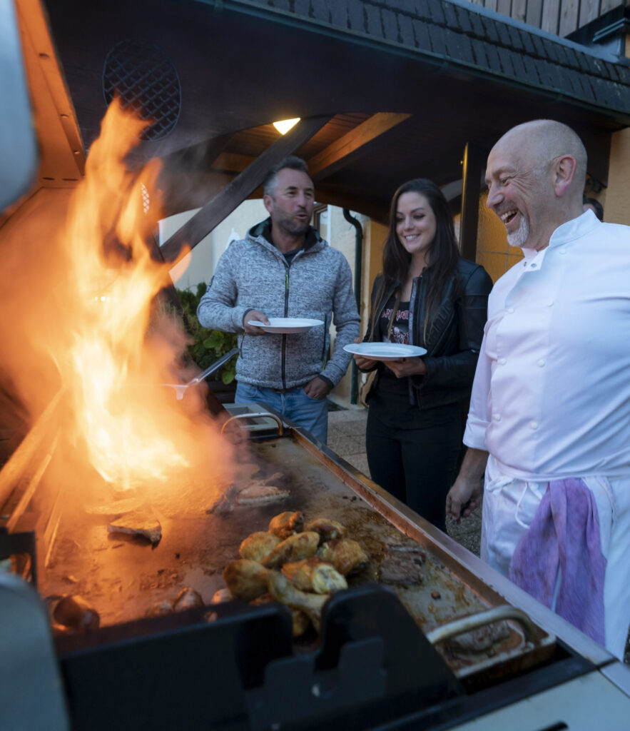 Ein Mann und eine Frau stehen lächelnd neben einem lachenden Koch, der Essen auf einem großen, flammenden Grill im Freien zubereitet.