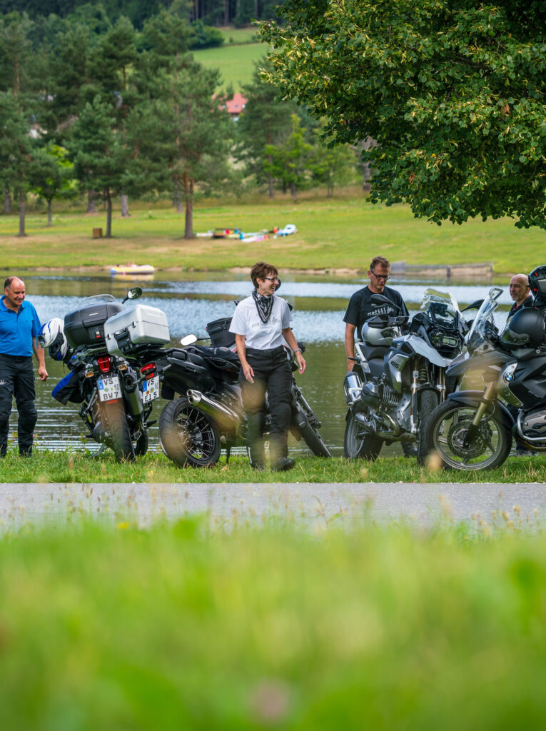 Gruppe von Menschen steht neben Motorrädern an einem Seeufer mit Bäumen und einer Wiese im Hintergrund.