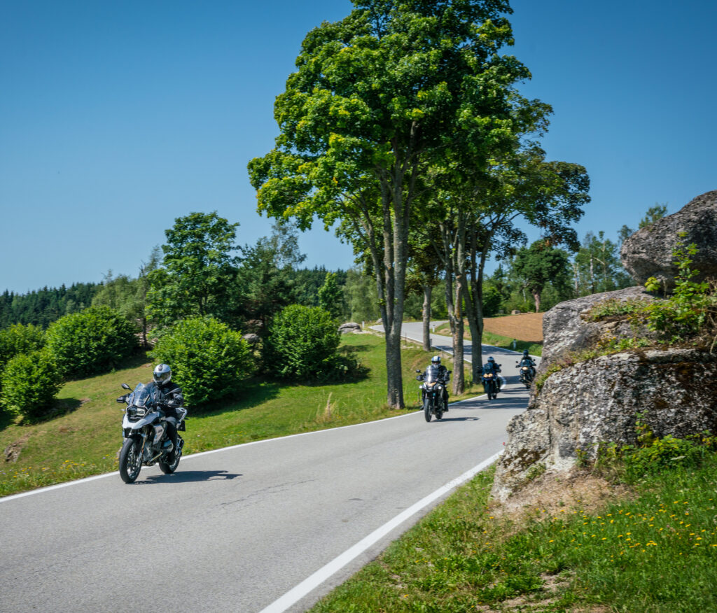 Motorradfahrer auf einer kurvigen Landstraße an einem sonnigen Tag, umgeben von grüner Landschaft und Felsbrocken.