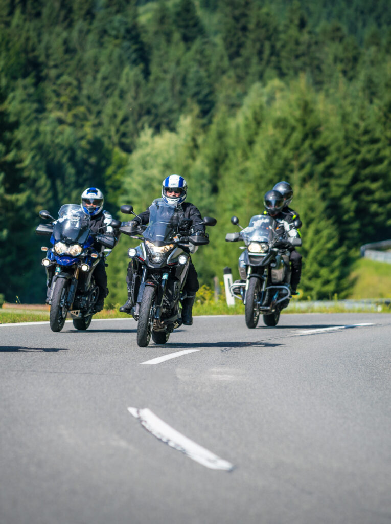 Drei Motorradfahrer fahren auf einer kurvigen Landstraße in einer bewaldeten Gegend.