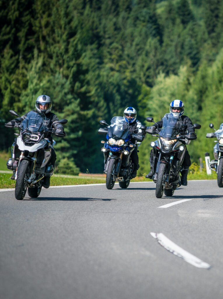Drei Motorradfahrer in Schutzkleidung fahren auf einer Landstraße, umgeben von grünen Bäumen.