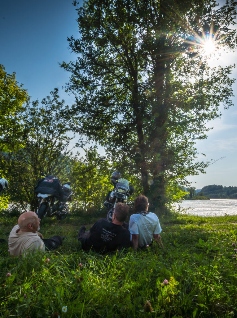 Gruppe von Menschen entspannt sich im Gras unter einem Baum am Ufer eines Flusses, mit Sonnenstrahlen, die durch die Blätter scheinen.