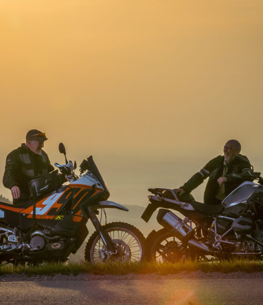 Zwei Motorradfahrer unterhalten sich am Straßenrand bei Sonnenuntergang.