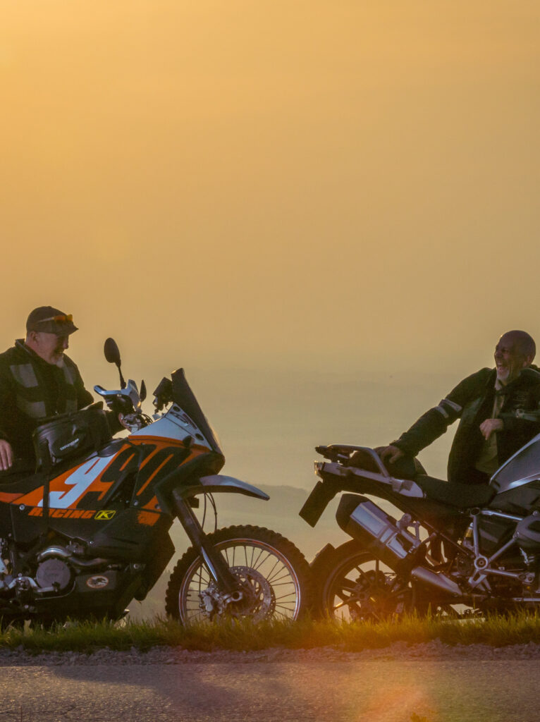 Zwei Motorradfahrer während des Sonnenuntergangs auf einer Anhöhe sitzend, unterhalten sich neben ihren parkenden Motorrädern.
