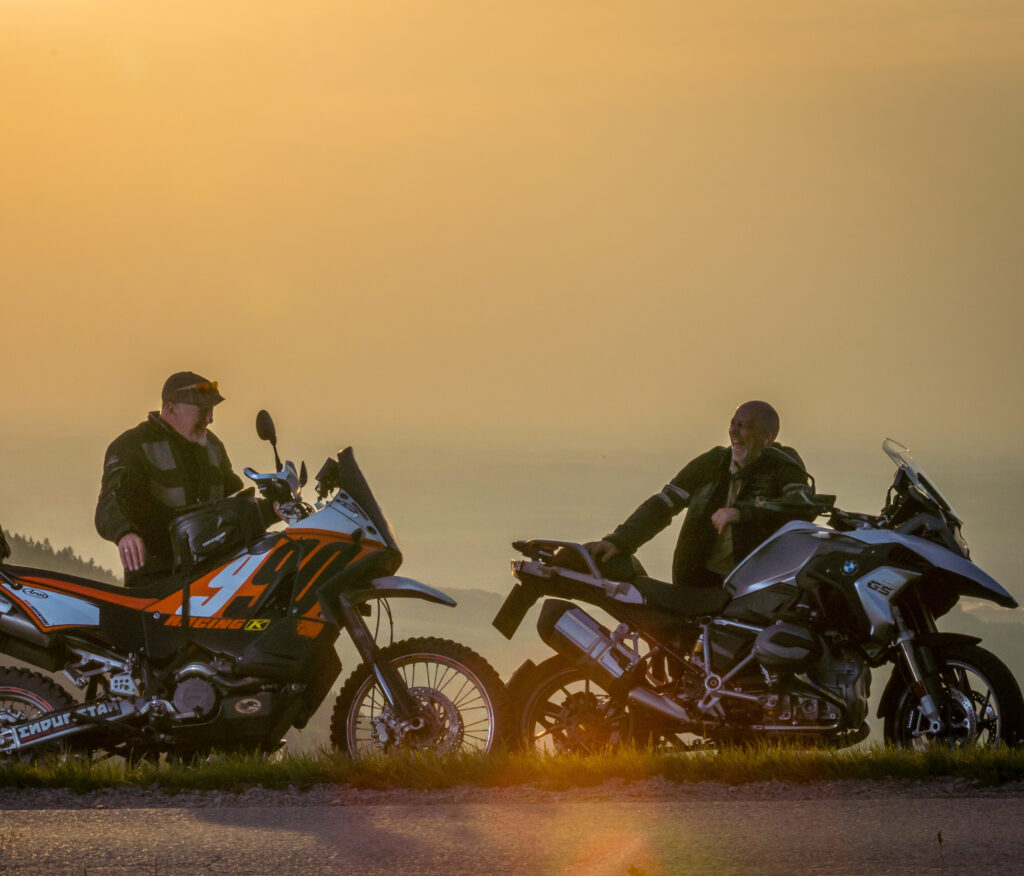 Zwei Personen sitzen auf Motorrädern und unterhalten sich bei Sonnenuntergang auf einem Hügel.