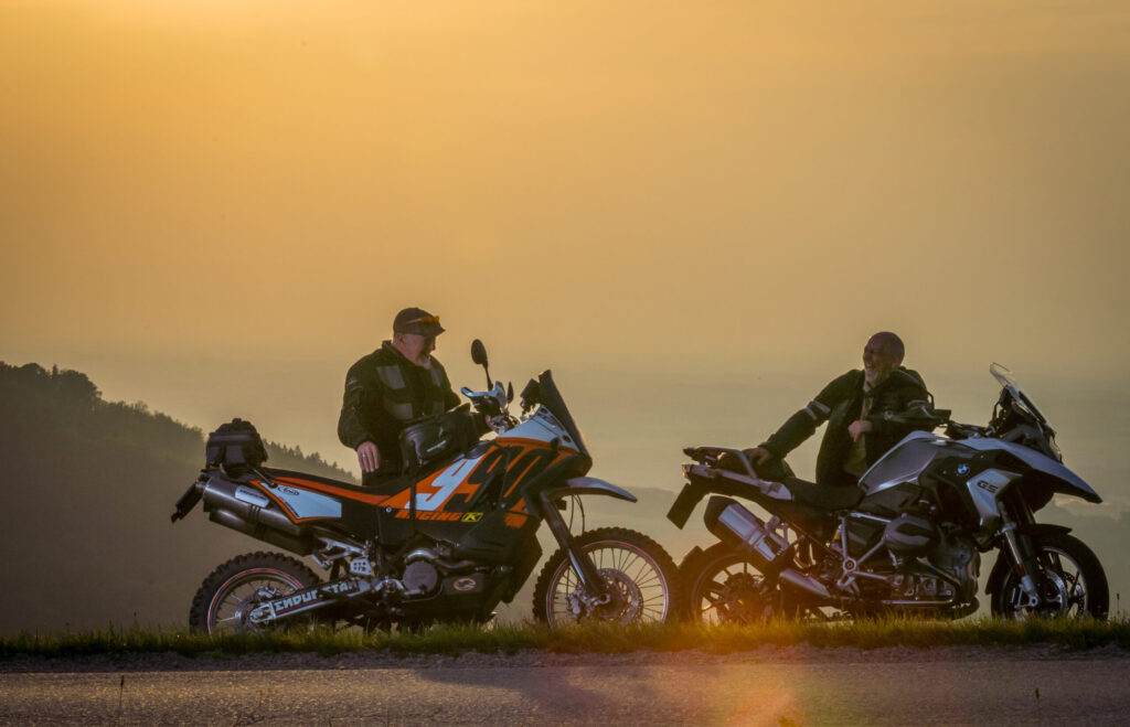Zwei Motorradfahrer sitzen bei Sonnenuntergang auf ihren Motorrädern an einem Aussichtspunkt und unterhalten sich.