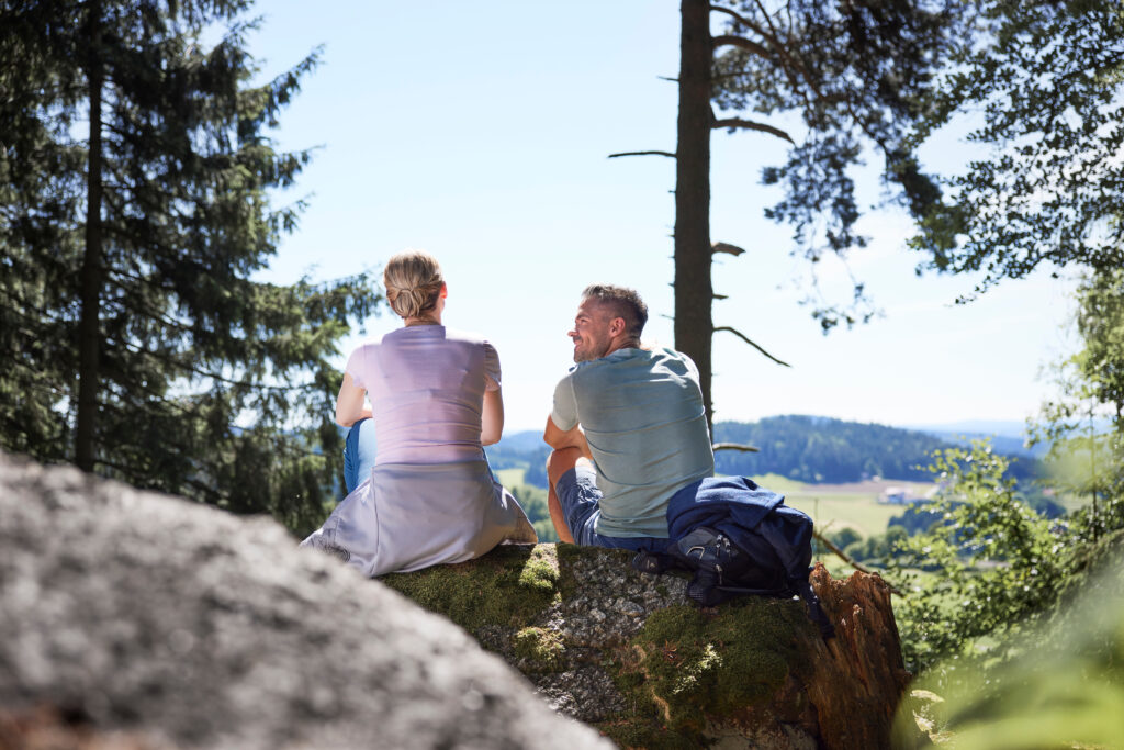 Ein Paar sitzt auf einem Felsen im Wald und blickt auf eine hügelige Landschaft.