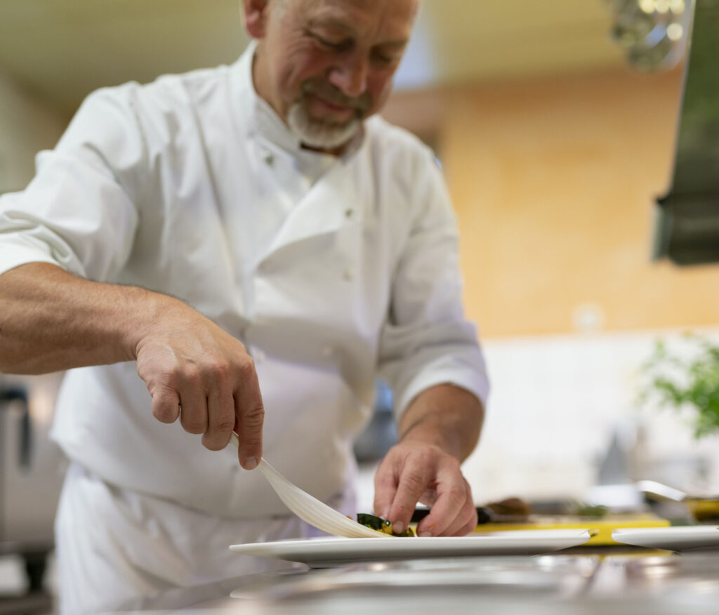 Ein erfahrener Chefkoch in weißer Uniform richtet sorgfältig ein Gericht auf einem weißen Teller in einer professionellen Küche an.