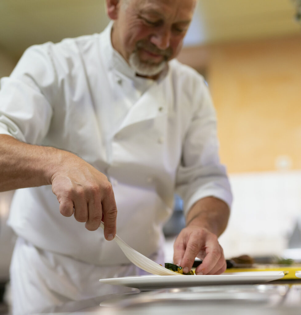 Ein älterer Koch in einer Küche, der sorgfältig ein feines Gericht auf einem weißen Teller anrichtet.