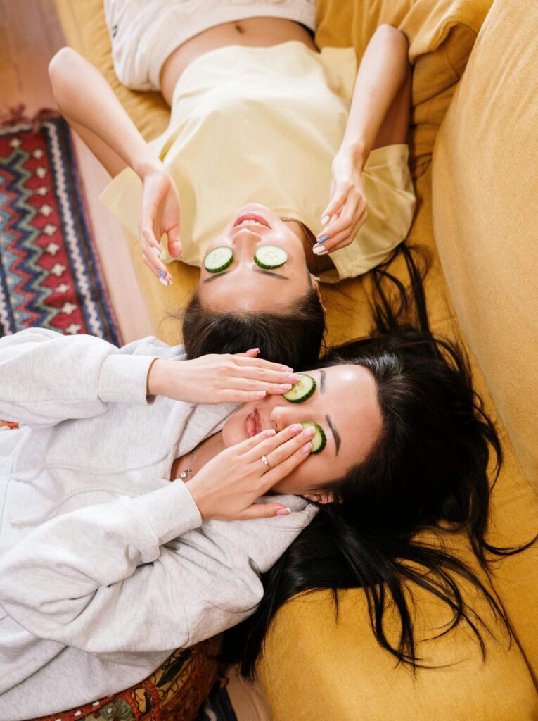 Zwei Frauen entspannen auf einem gelben Sofa, Gesichter mit Gurkenscheiben bedeckt, während sie eine Wellnessbehandlung genießen.