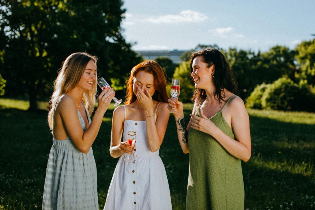 Drei Frauen stoßen mit Weingläsern an und lächeln sich in einem sonnigen Park an.