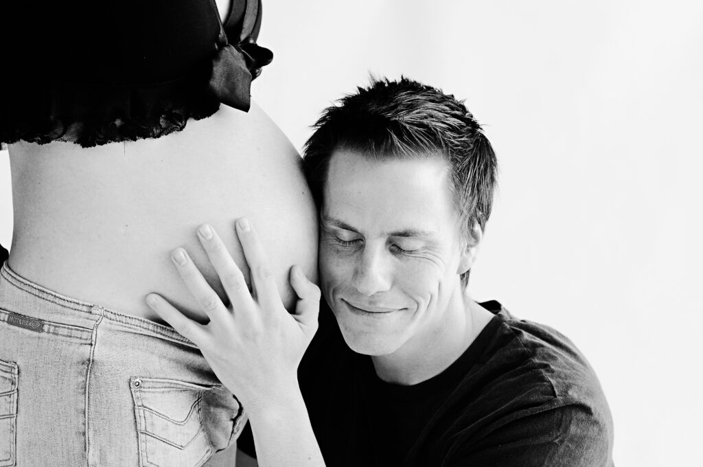 Ein Mann lächelt und legt seine Hand auf den schwangeren Bauch einer Frau. Sie stehen auf einem hellen Hintergrund.