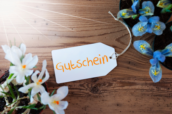 Ein Schild mit der Aufschrift "Gutschein" liegt auf einem Holztisch neben einer Pflanze mit blauen Blüten.